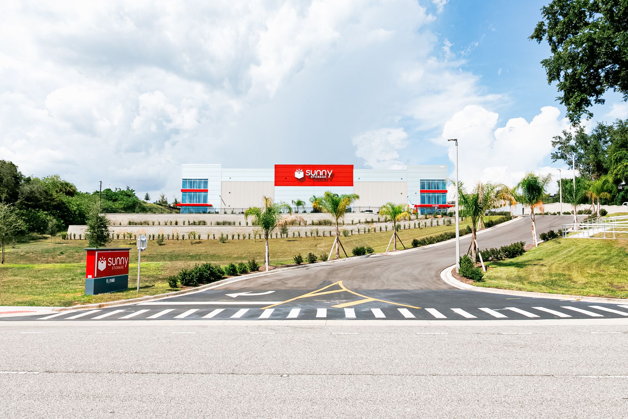Sunny Storage self storage building on Hooks St. in Clermont, Florida, with the red Sunny Storage sign on a white and grey facade, palm trees and a blue sky.